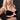 Person wearing a black bra top with a chain detail and pink Vixen Female Chest Harness against a dark background.