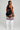 A woman stands in profile wearing a black sports bra and shorts with white side panels, paired with white knee-high socks featuring navy and red stripes, against a light gray studio background.