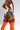 A model wearing a white bodysuit and bright orange thigh-high socks with black stripes, shown from a side-rear angle against a light gray studio background.