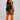 A woman modeling a black athletic crop top and shorts set with white piping, paired with bright orange over-the-knee socks featuring black stripes, shown from a rear three-quarter angle against a light gray studio background.