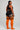 A woman modeling a black athletic crop top and shorts set with white piping, paired with bright orange over-the-knee socks featuring black stripes, shown from a rear three-quarter angle against a light gray studio background.