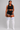 A woman modeling a black athletic crop top and matching black shorts with white side stripes, paired with white thigh-high socks featuring pink and blue stripes near the top, against a light gray studio background.