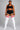 A plus-size model stands against a gray studio background wearing a black sports bra and shorts with white side stripes, paired with white thigh-high socks featuring red stripes near the top.
