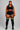 A plus-size model stands against a solid gray studio background wearing a matching black activewear set consisting of a sports bra, high-waisted shorts with white piping, and black over-the-knee socks featuring yellow, orange, and teal stripes near the top of the legs.