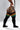A model wearing black ribbed thigh-high socks with horizontal stripes in green, yellow, orange, and white, paired with a white bodysuit. The photo is taken from a side-rear angle against a light gray studio background.