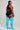A woman stands in profile against a light gray studio background, wearing a black sports bra and black athletic shorts with white side trim, paired with bright turquoise ribbed knee-high socks.