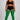 A model stands against a light gray studio background wearing a black sports bra and shorts with white side stripes, paired with bright green ribbed over-the-knee socks.