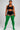 A model stands against a light gray studio background wearing a black sports bra and shorts with white side stripes, paired with bright green ribbed over-the-knee socks.