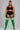 A plus-size model stands against a solid gray studio background wearing a black sports bra, black athletic shorts with white side stripes, and bright green thigh-high socks with white stripes near the top.