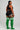 A woman stands in profile wearing a black sports bra and shorts with white side stripes, paired with bright green over-the-knee socks featuring white horizontal stripes near the top. She is photographed against a plain light gray studio background.