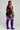 A woman stands in profile wearing a black sports bra and shorts with white side stripes, paired with purple over-the-knee socks featuring white horizontal stripes near the top.