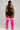 A woman shown from the back wearing a black sports bra and matching black athletic shorts with white trim, paired with bright pink over-the-knee socks featuring three white stripes near the top, against a light gray studio background.