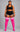 A plus-size model stands against a gray studio background wearing a black sports bra and shorts with white side stripes, paired with bright pink over-the-knee socks featuring white horizontal stripes near the top.