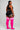 A woman stands in profile against a light gray studio background, wearing a black athletic crop top and shorts with white trim, paired with bright pink over-the-knee socks featuring three white horizontal stripes near the top.