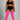 A plus-size model stands against a neutral gray studio background wearing a black sports bra and shorts with white side stripes, paired with bright pink over-the-knee socks.
