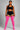 A plus-size model stands against a neutral gray studio background wearing a black sports bra and shorts with white side stripes, paired with bright pink over-the-knee socks.