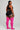 A woman stands in profile against a light gray studio background, wearing a black sports bra and black athletic shorts with white side trim, paired with bright pink ribbed over-the-knee socks.
