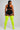 A woman shown from the back wearing a black sports bra and black shorts with white trim, paired with bright neon yellow thigh-high socks, against a light gray studio background.