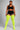 A model wearing a black sports bra and shorts with white side stripes, paired with bright neon yellow thigh-high socks, against a solid gray studio background.