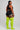 A woman stands in profile wearing a black sports bra and black athletic shorts with white side trim, paired with bright neon yellow-green ribbed over-the-knee socks, against a light gray studio background.