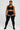 A plus-size model stands against a light gray studio background wearing a black sports bra, black shorts with white side stripes, and black thigh-high socks.