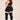 A woman with long red hair is shown from the back wearing a black ribbed crop top, black ribbed shorts, and black ribbed thigh-high socks against a plain beige studio background.