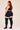 A woman with long red hair is shown from the back wearing a black ribbed crop top, black ribbed shorts, and black ribbed thigh-high socks against a plain beige studio background.