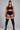 A plus-size model stands against a neutral gray studio background wearing a black athletic crop top, black shorts with white side stripes, sheer black thigh-high stockings with white stripes near the top, and black tights.