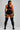 A woman shown from the back wearing a black sports bra and matching high-waisted shorts with white side trim, paired with black thigh-high socks featuring orange stripes near the top. She stands against a light gray studio background.