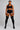 A model stands against a light gray studio background wearing a black sports bra, black athletic shorts with white side stripes, and black thigh-high socks with orange stripes near the top.