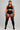 A plus-size model stands against a light gray studio background wearing a black athletic halter crop top, black shorts with white side stripes, and black thigh-high socks with red stripes near the top.