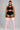 A plus-size model stands against a light gray studio background wearing a black sports bra and matching black athletic shorts with white side stripes, paired with light pink ribbed knee-high socks featuring white stripes near the top.