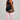 A woman stands in profile against a light gray studio background, wearing a black sports bra and black athletic shorts with white side stripes, paired with light pink ribbed knee-high socks featuring white horizontal stripes near the top.