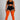 A plus-size model stands against a light gray studio background wearing a black sports bra and shorts, sheer black thigh-high stockings, and bright orange over-the-knee socks.