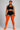 A plus-size model stands against a light gray studio background wearing a black sports bra and shorts, sheer black thigh-high stockings, and bright orange over-the-knee socks.