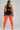 A woman shown from the back wearing a black sports bra and black athletic shorts with white trim, paired with bright orange ribbed thigh-high socks, against a light gray studio background.
