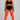 A model stands against a light gray studio background wearing a black sports bra and shorts with white trim, paired with bright orange ribbed over-the-knee socks.