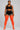 A model stands against a light gray studio background wearing a black sports bra and shorts with white trim, paired with bright orange ribbed over-the-knee socks.