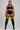 A plus-size model stands against a light gray studio background wearing a black sports bra, black athletic shorts with white side stripes, and black thigh-high socks with yellow stripes near the top.