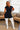 A woman modeling a black short-sleeve zip-front bodysuit with matching black shorts and white thigh-high socks in a bright living room setting.